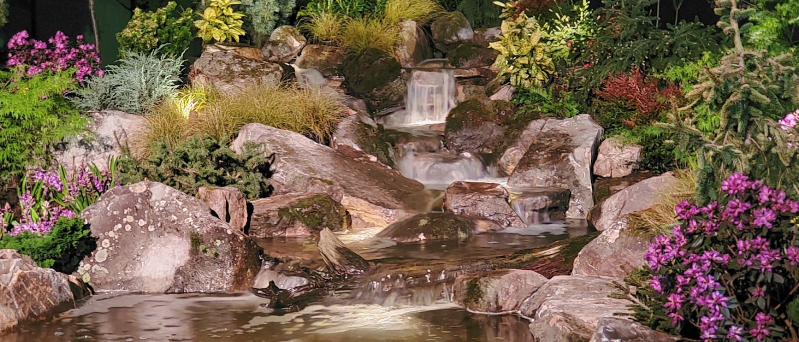 Water cascading over stone through a lush, lit planting bed