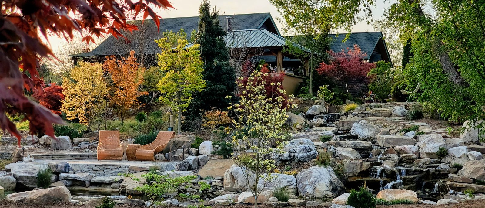 A Japanese maple frames a multi-tier boulder waterfall, patio, and timber-and-stone home
