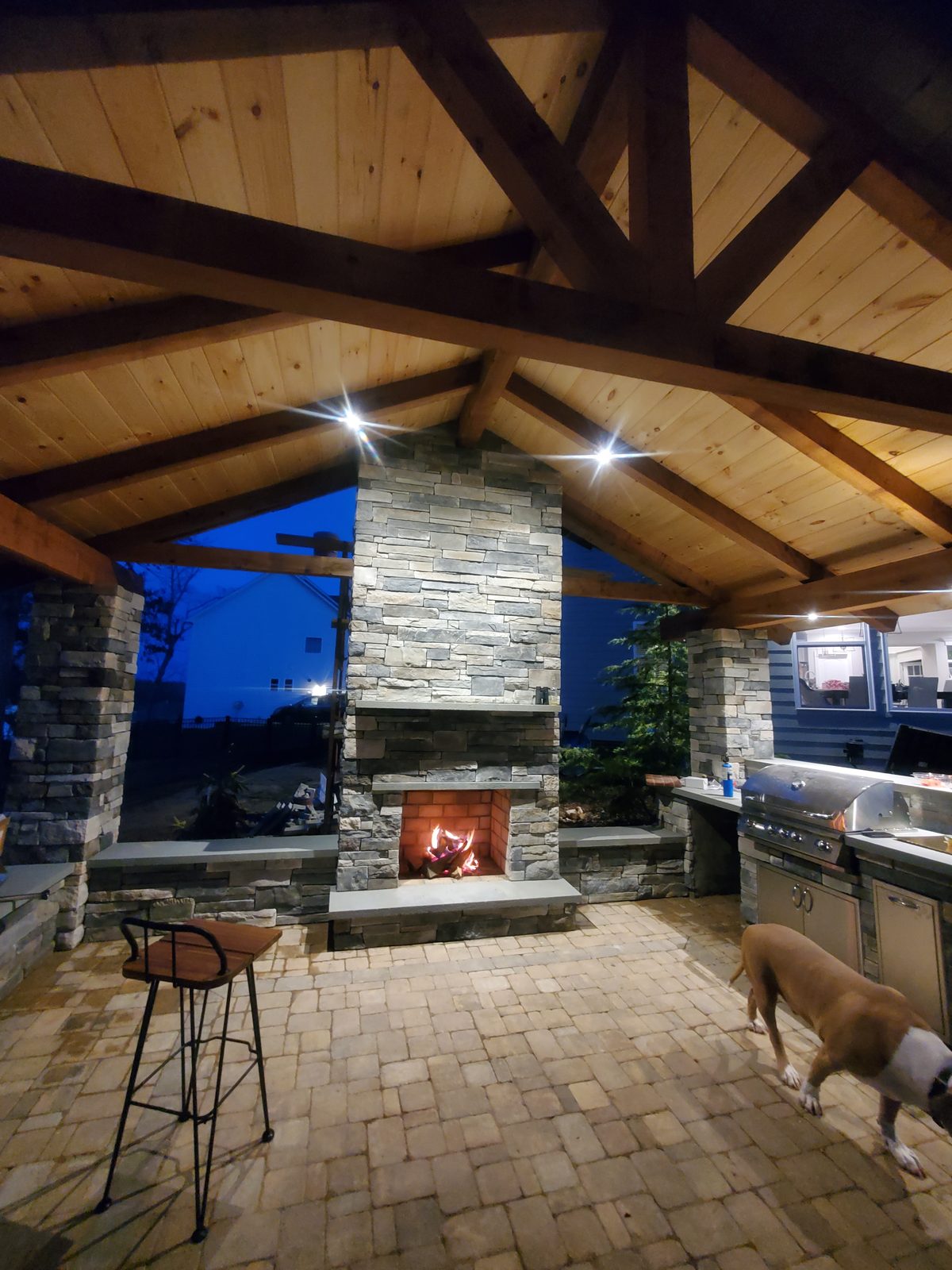 Pavilion interior with stone fireplace and outdoor kitchen, dusk