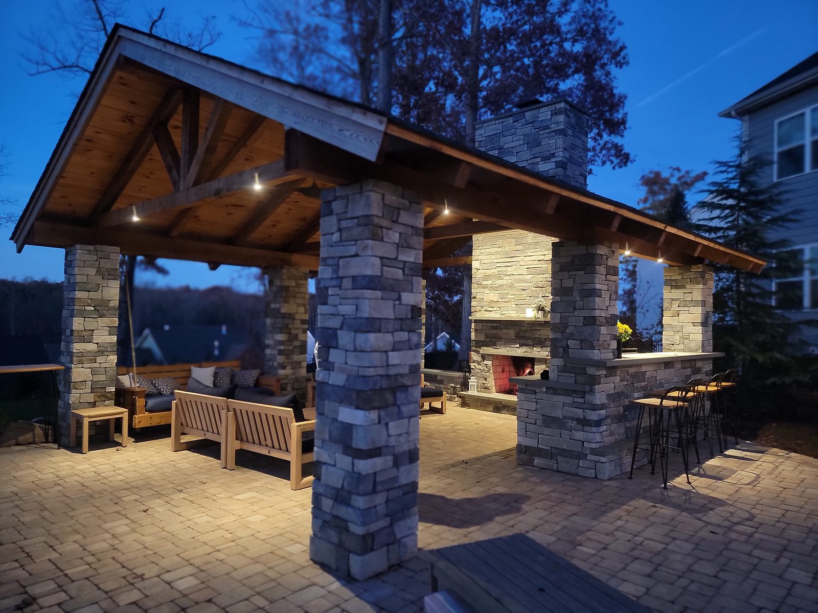 Timber-frame pavilion with stacked-stone fireplace and outdoor bar, lit at dusk