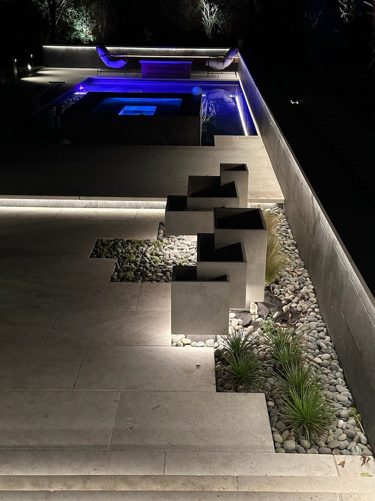 The floating limestone planter steps underlit at night, with the pool and spa glowing blue behind them