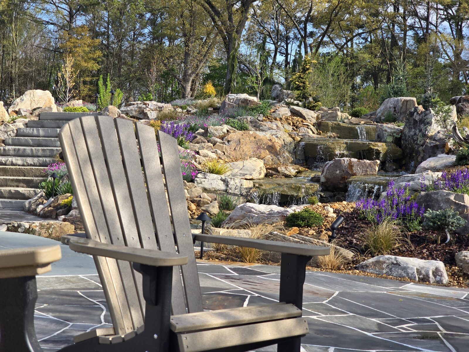 An Adirondack chair on the patio with the boulder waterfall and spring perennial garden beyond