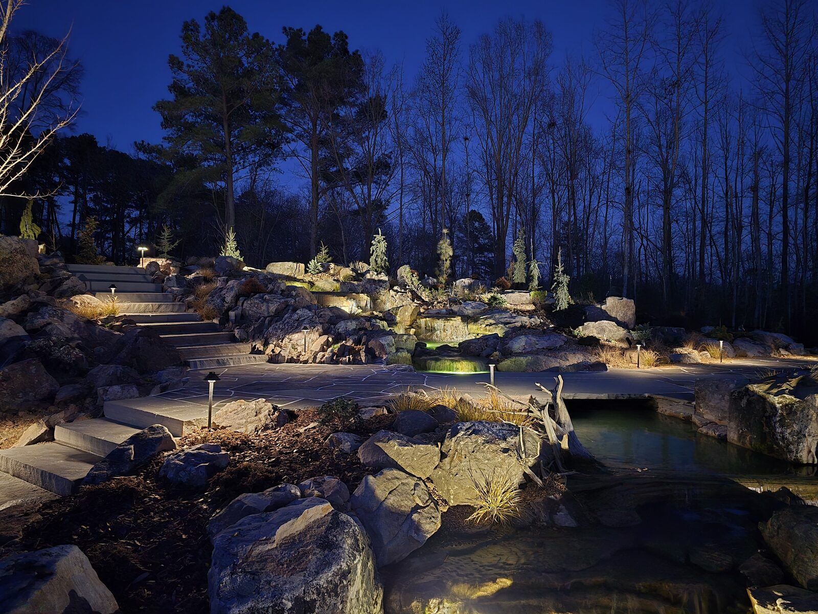 The multi-tier boulder waterfall and stone steps illuminated at night, showing the layered landscape lighting design