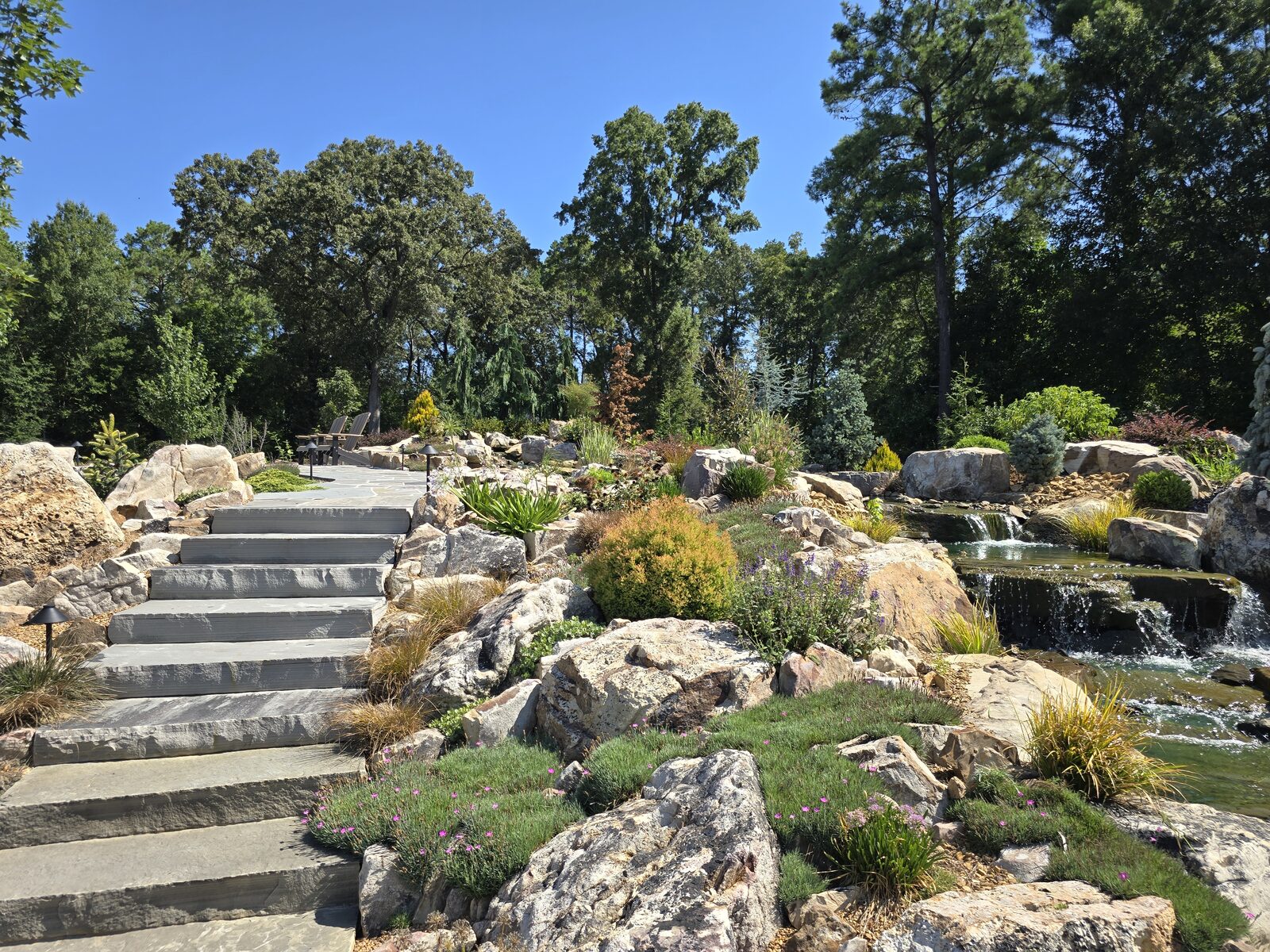 Wide summer view of the finished stone steps, patio, and boulder waterfall with seating overlooking the water