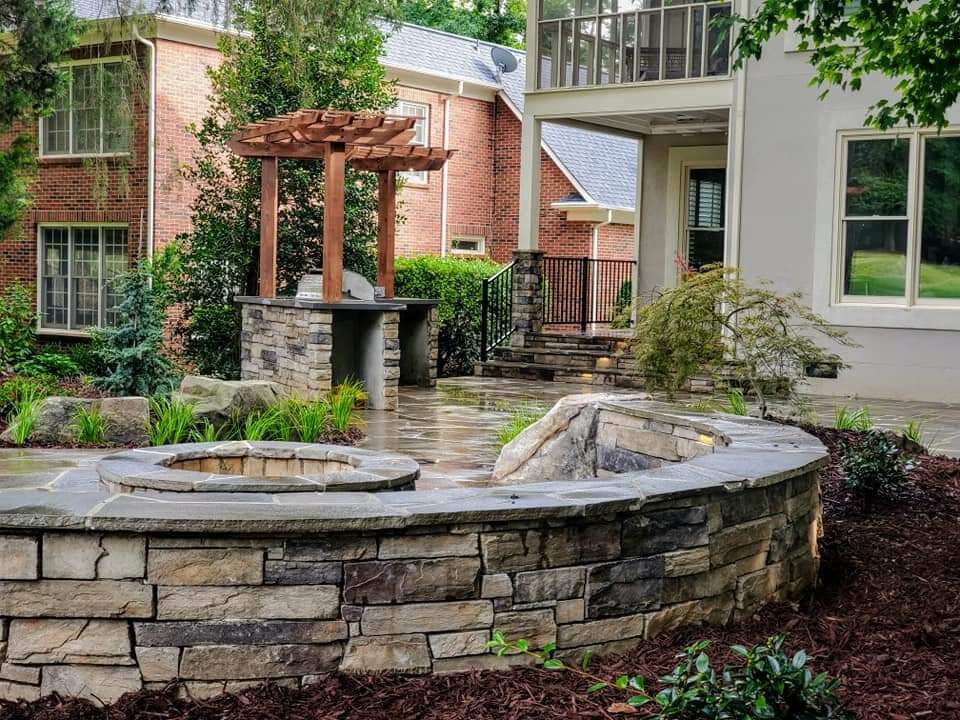 A circular flagstone patio with a stacked-stone seat wall, in-ground fire pit, and timber pergola over an outdoor kitchen