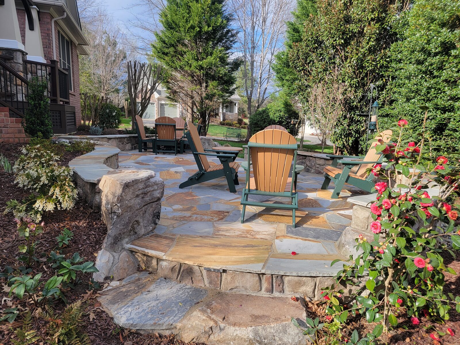 A curved flagstone patio with a stone seat wall, fire pit, and Adirondack chairs surrounded by garden planting