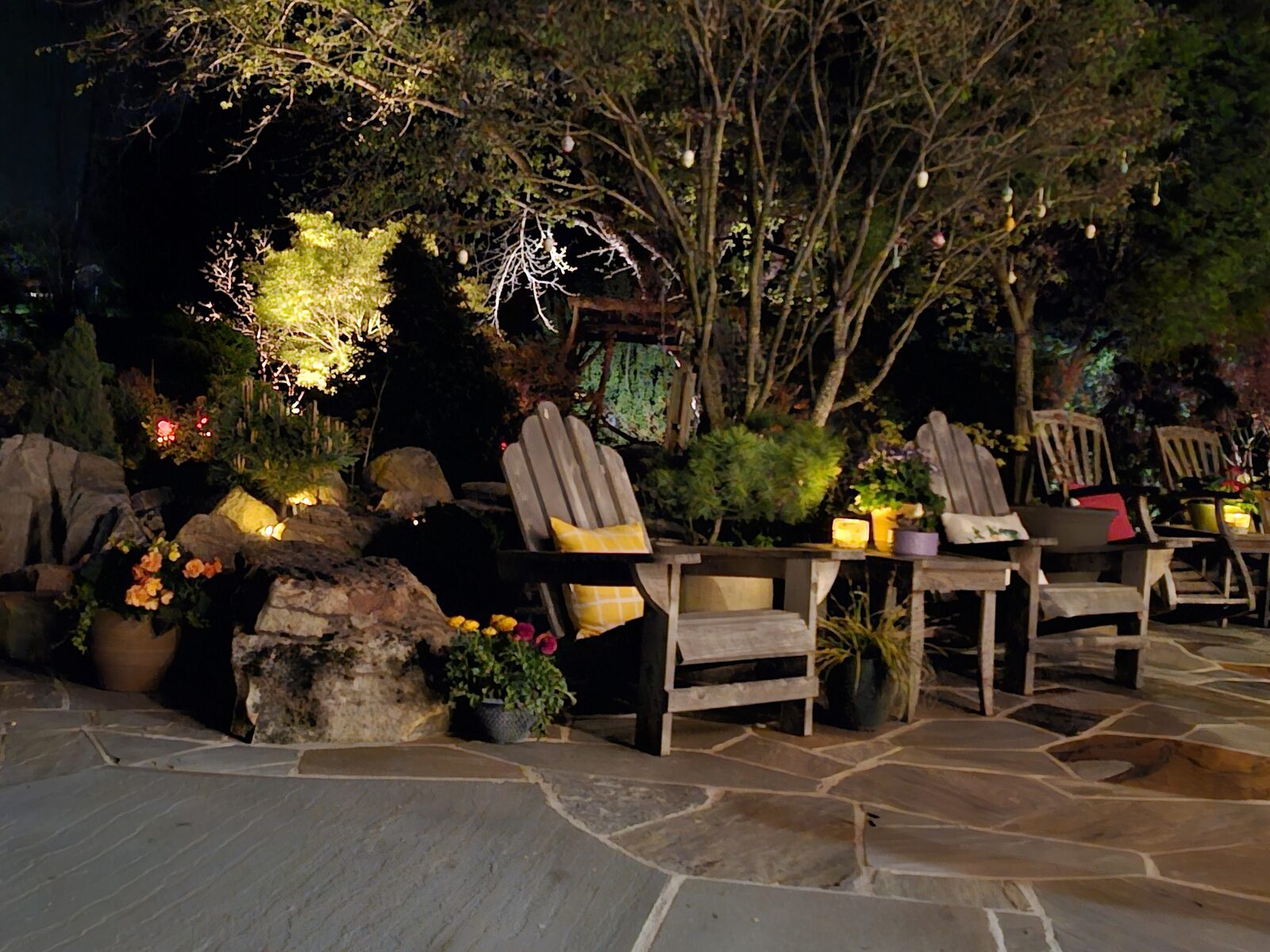 A flagstone patio at night, with garden lighting illuminating boulders, planting, and wooden chairs