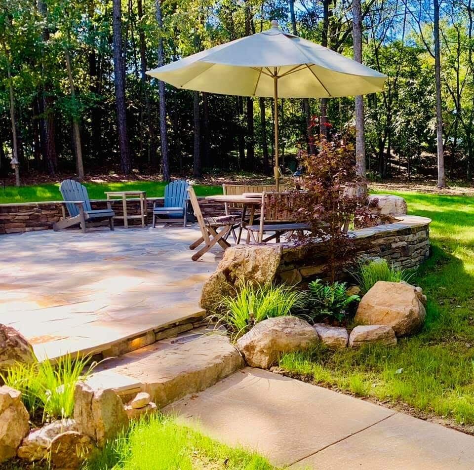 A raised flagstone patio with a stacked-stone retaining wall, dining table, and umbrella, set into a wooded lawn