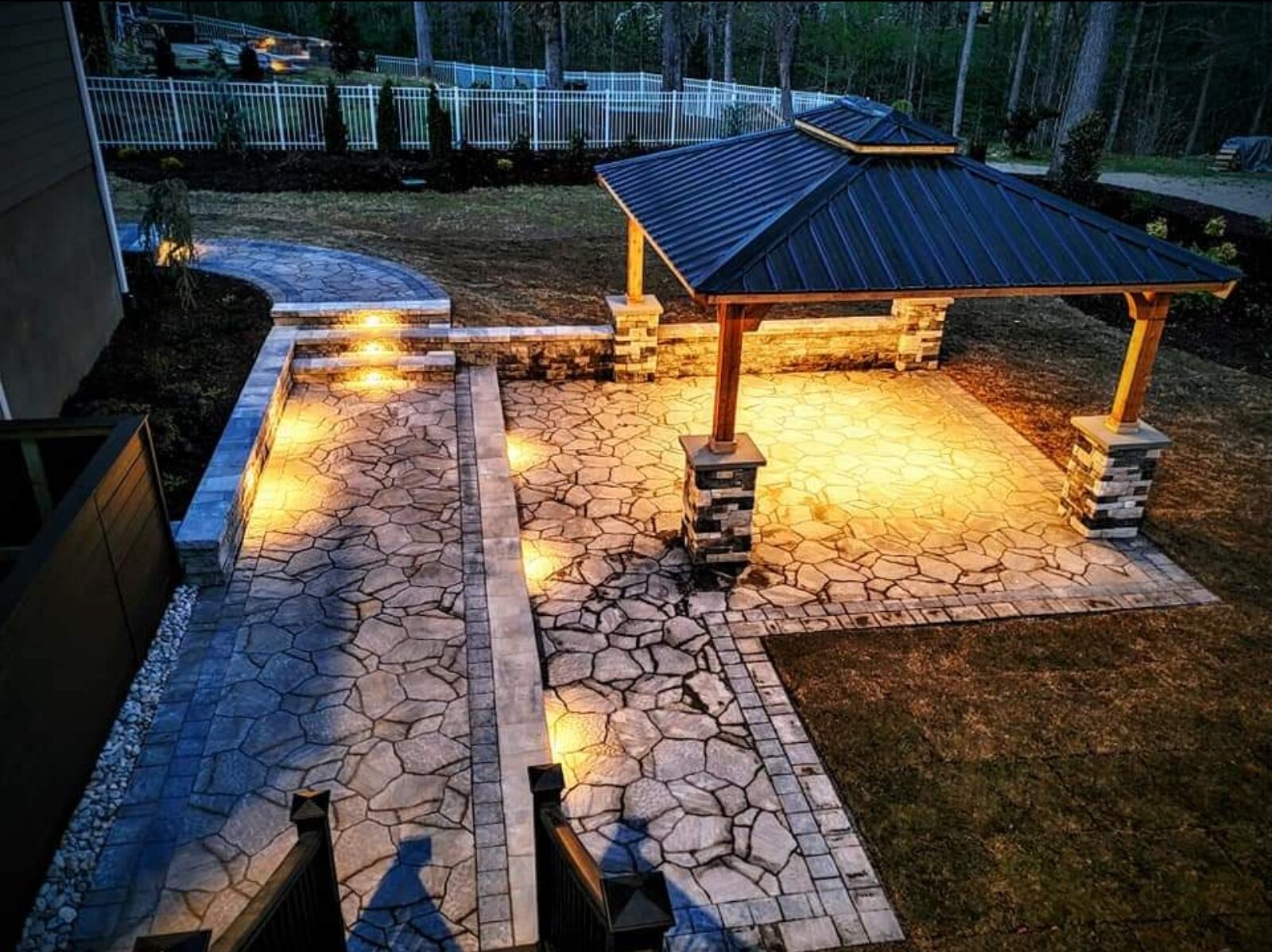 Aerial view of a stamped-concrete patio and walkway leading to a timber pavilion, lit at dusk