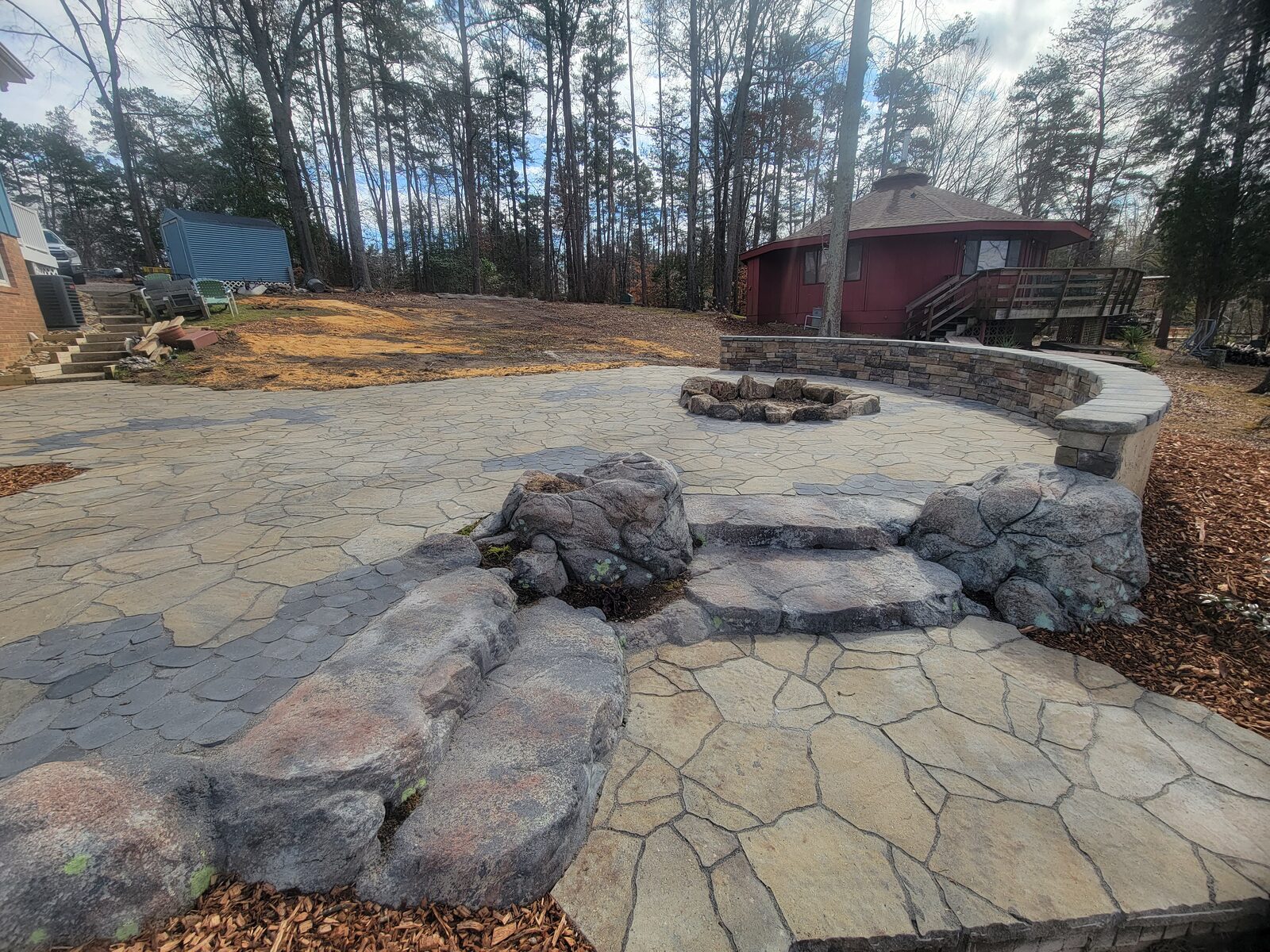 A circular flagstone patio with a fire pit and stacked-stone seat wall, surrounded by boulders and a cabin beyond