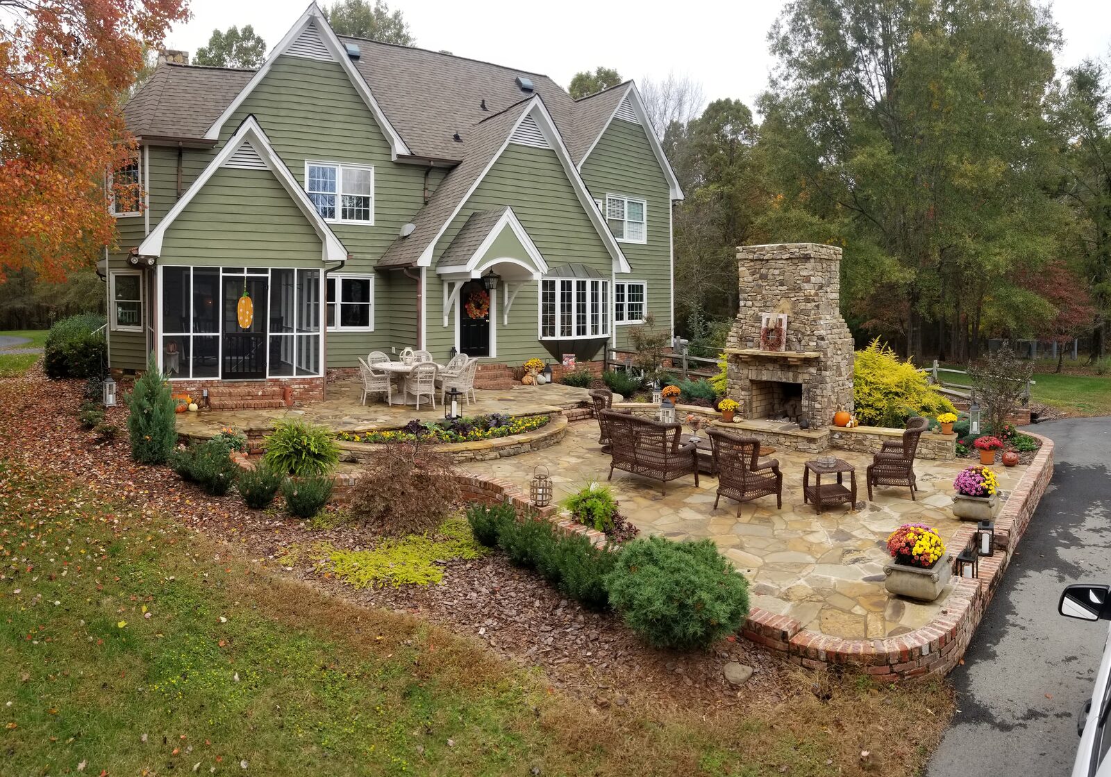 A large stone patio with wicker seating and a tall stacked-stone outdoor fireplace, surrounded by fall foliage