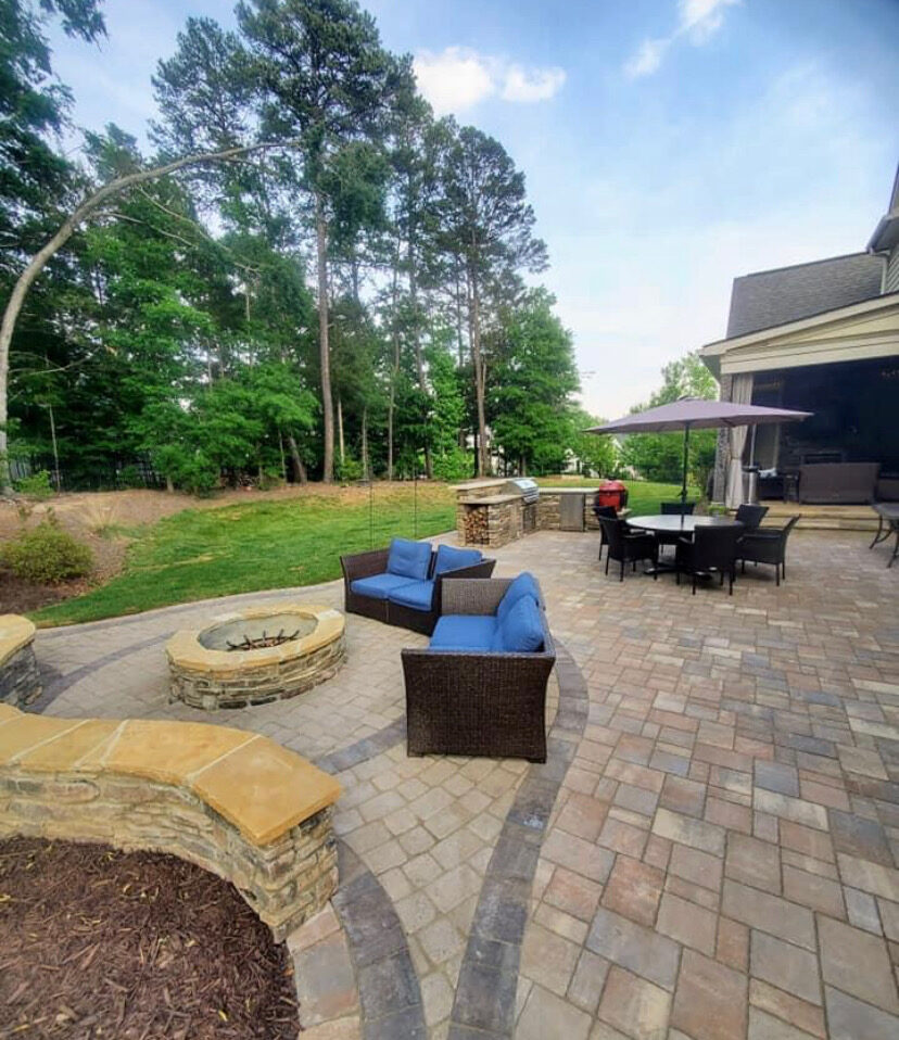 A modern paver patio with wicker furniture, a built-in fire pit, an outdoor kitchen, and a market umbrella over a dining table