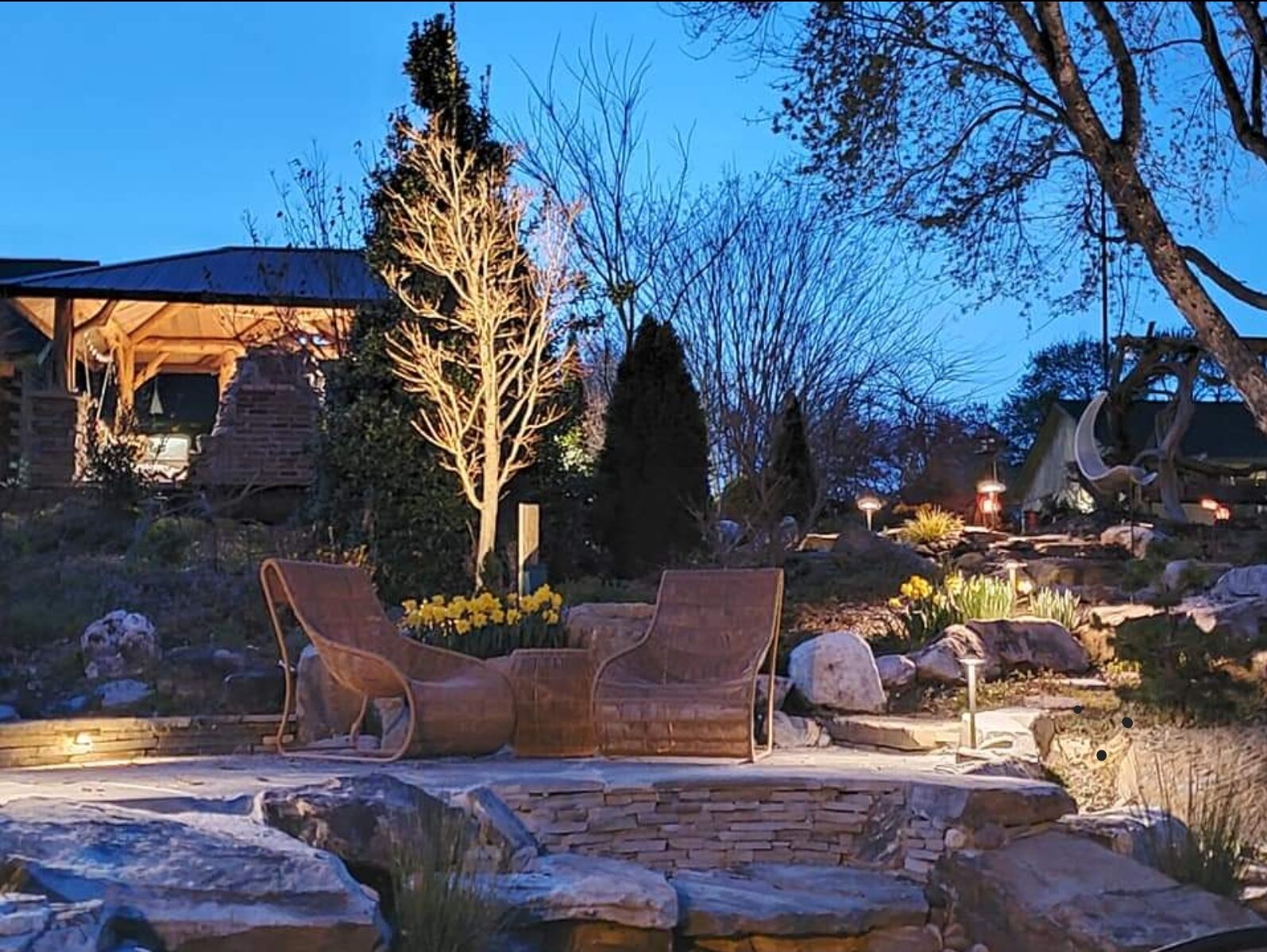 Sculptural metal lounge chairs beside a stone fire pit at dusk, with landscape lighting through the garden beyond