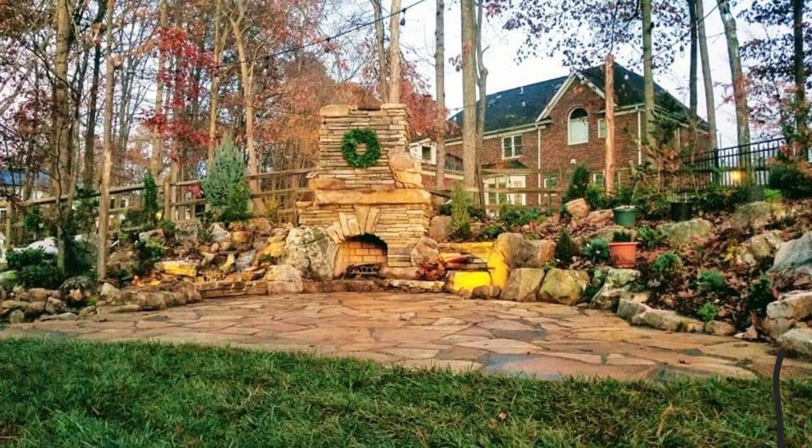 A tall stacked-stone outdoor fireplace on a flagstone patio, lit at dusk with string lights overhead