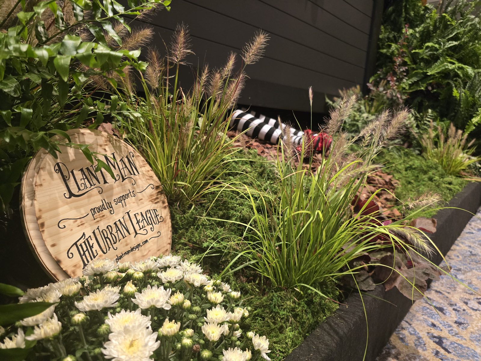 A wooden sign reading Plant Man proudly supports The Urban League, set among ornamental grasses and mums