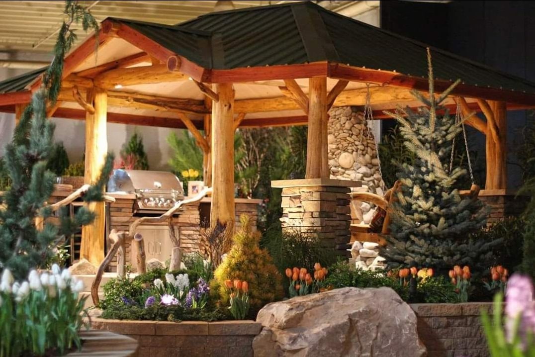 A temporary timber pavilion with a stacked river-rock chimney and outdoor kitchen, surrounded by spring tulips and a boulder planting bed
